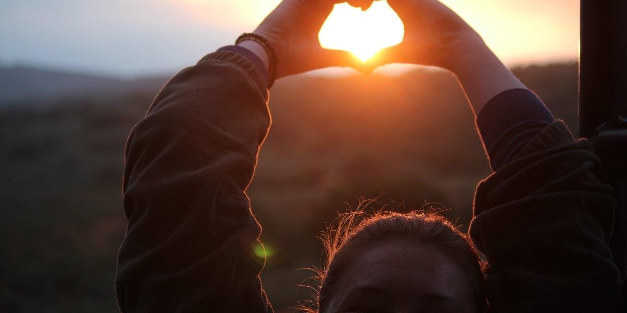 Aufnahme einer Field Guide-Studentin im suedafrikanischen Ostkap, die mit ihren Haenden ein Herz vor der untergehenden Sonne formt