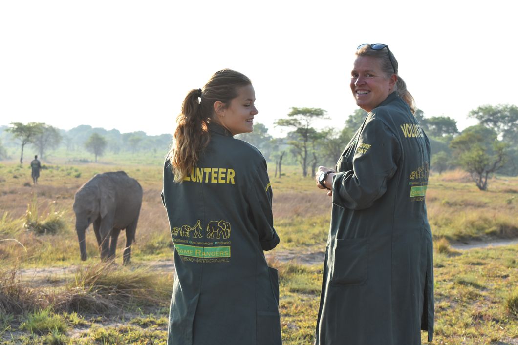 Zwei Volunteers des Elefantenprojekts in Sambia stehen zusammen und lachen; im Hintergrund laeuft ein Elefant