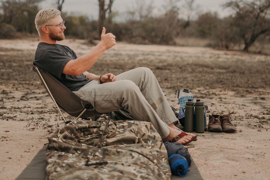 The outdoor night camp on a durable surface shows a ground tarp, with air mattress and sleeping bag. Behind it, a participant sits contentedly in his folding trail chair, with his equipment beside him.