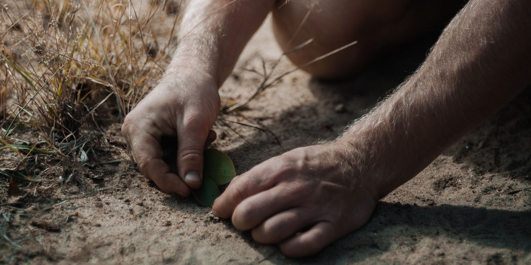 A close-up of someone kneeling and picking up something from the sandy ground with his hands.
