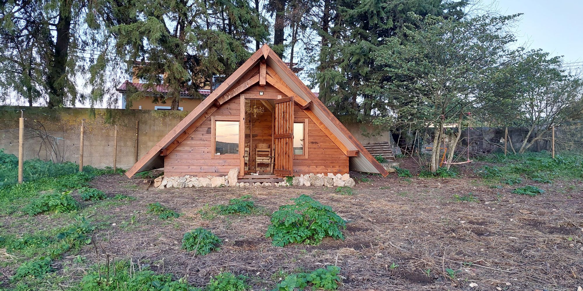 Aus ein paar Metern Entfernung ist das dreieckige, hölzerne Hühnerhaus vor einer Reihe aus Nadelbäumen zusehen.
