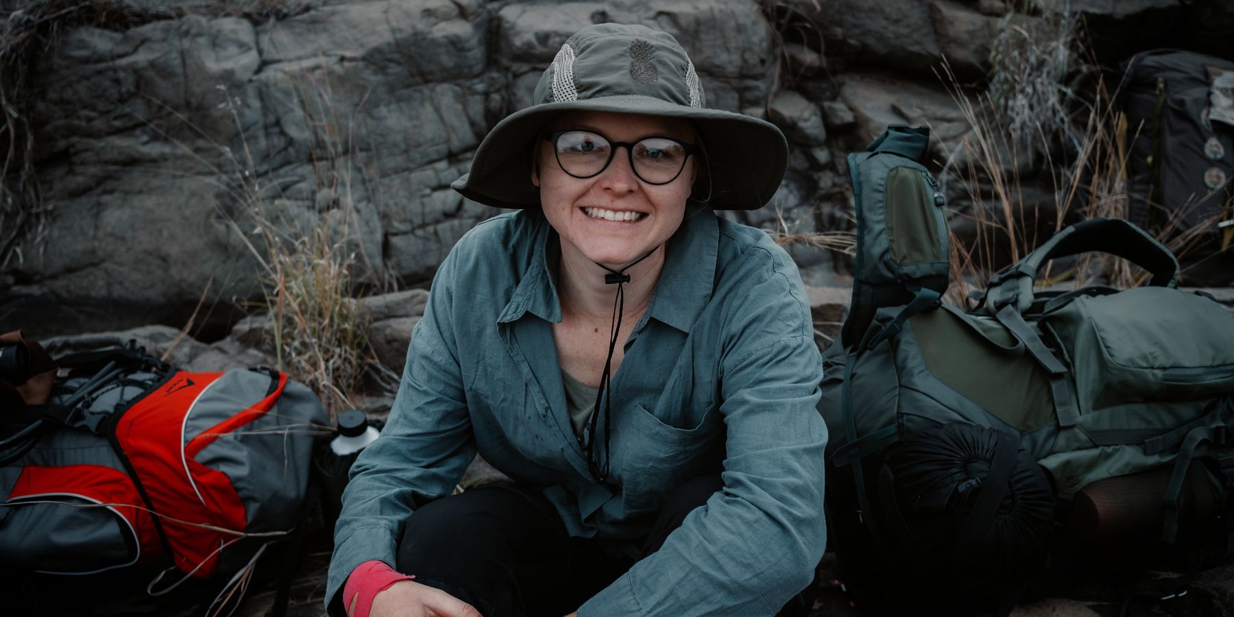 A happy EcoStar Nature Course participant sits on the ground in front of a rock face in South Africa, surrounded by her trail gear.