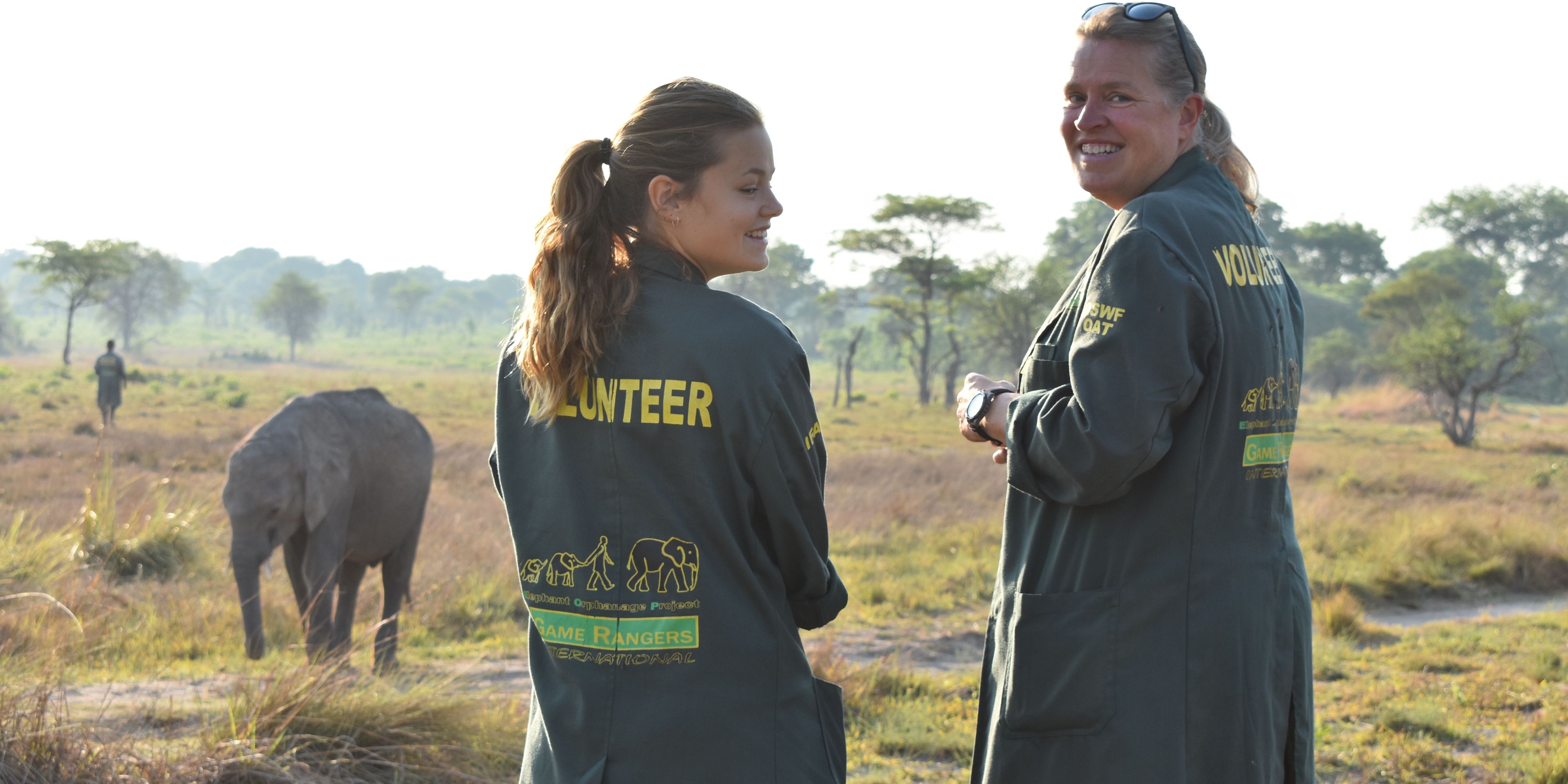 Zwei Volunteers des Elefantenprojekts in Sambia stehen zusammen und lachen; im Hintergrund laeuft ein Elefant