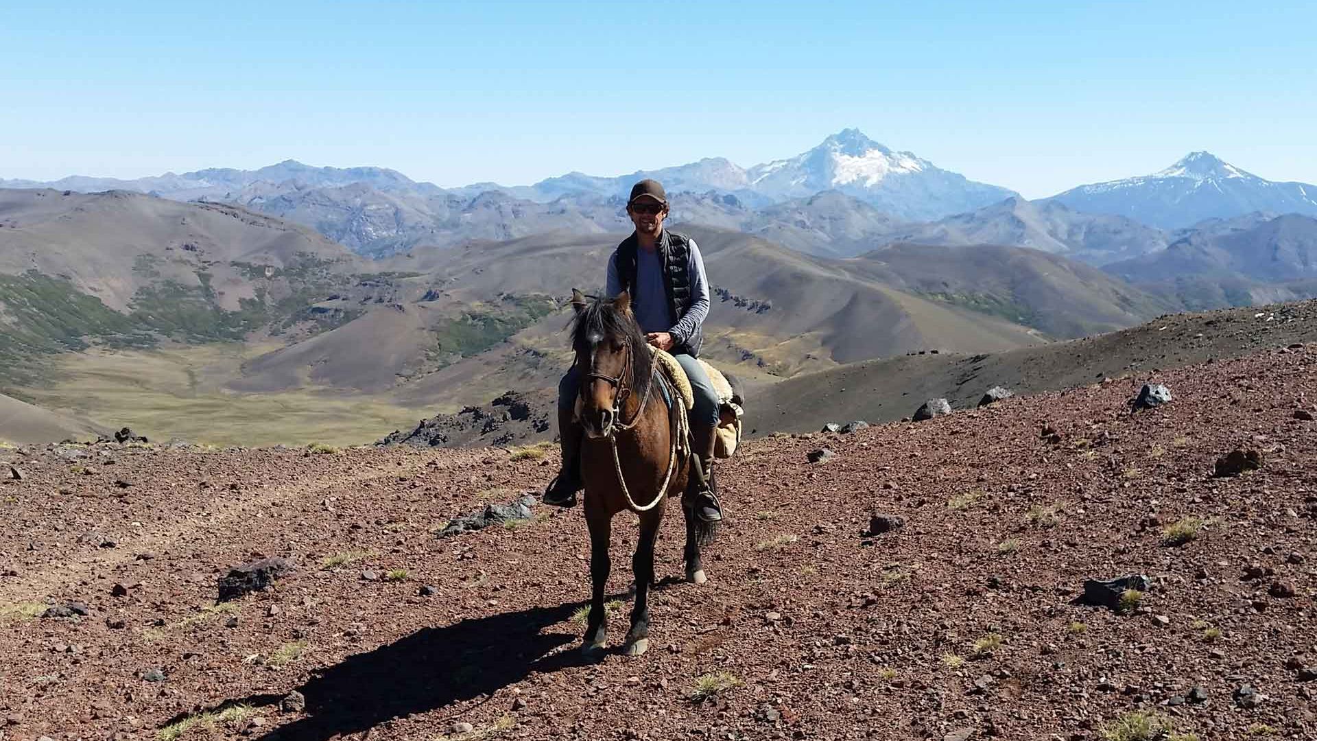 Ein Reiter sitzt auf einem Pferd und posiert fuer die Kamera vor einer Gebirgskette in den Anden