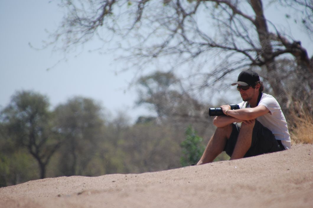 Ein Teilnehmer sitzt mit seiner Kamera in der Hand wartend auf einer Sandduene im afrikanischen Busch