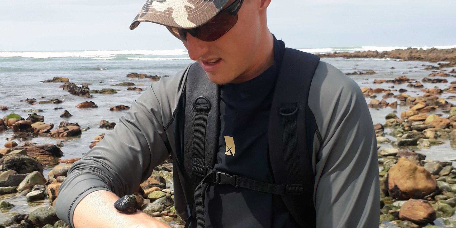 Marine Guide-Student steht an der Kueste Suedafrikas und beaeugt eine Schnecke, die auf seinem Unterarm sitzt