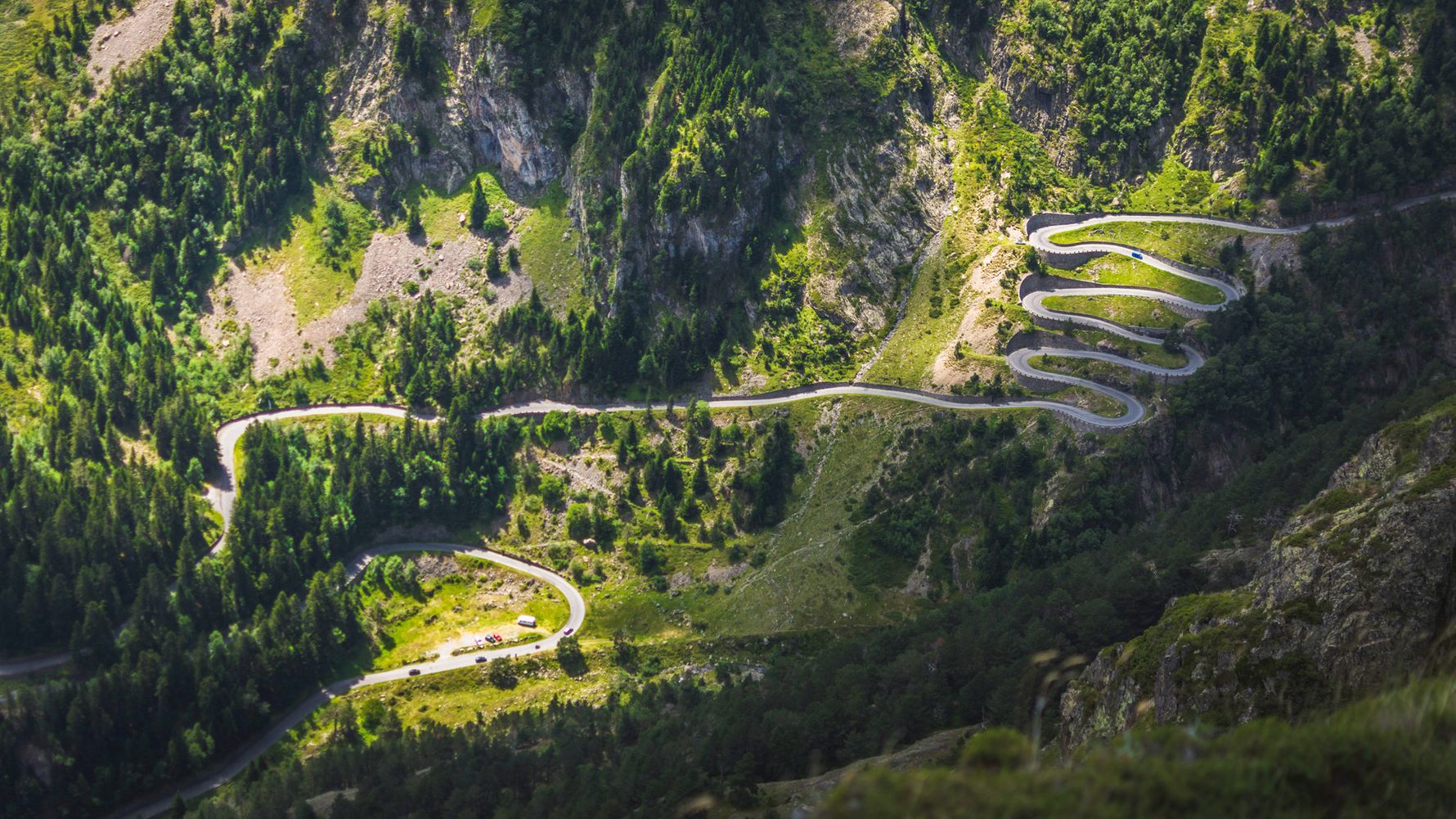 This valley in the Pyrenees is crossed by a winding road over serpentines and many trees green the area, which is only partially illuminated by sunlight on this cloudy day.