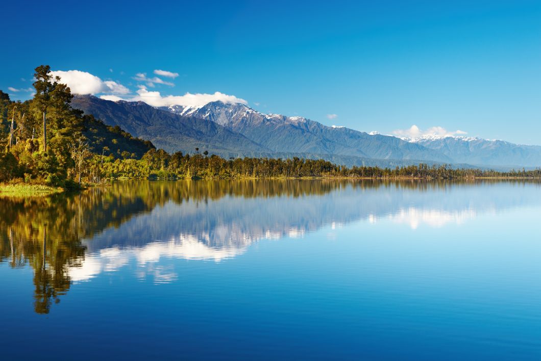 Blick auf einen See in Neuseeland mit dahinterliegendem Wald und Gebirgszuegen