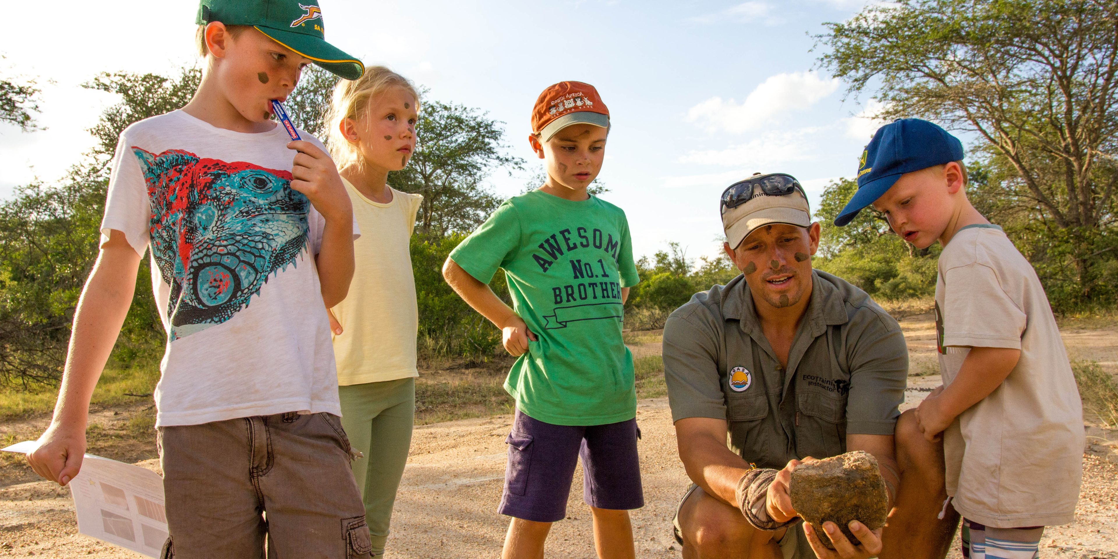 Vier Kinder stehen im Kreis um einen Guide, der ihnen das lokale Gestein waehrend einer Safari erklaert