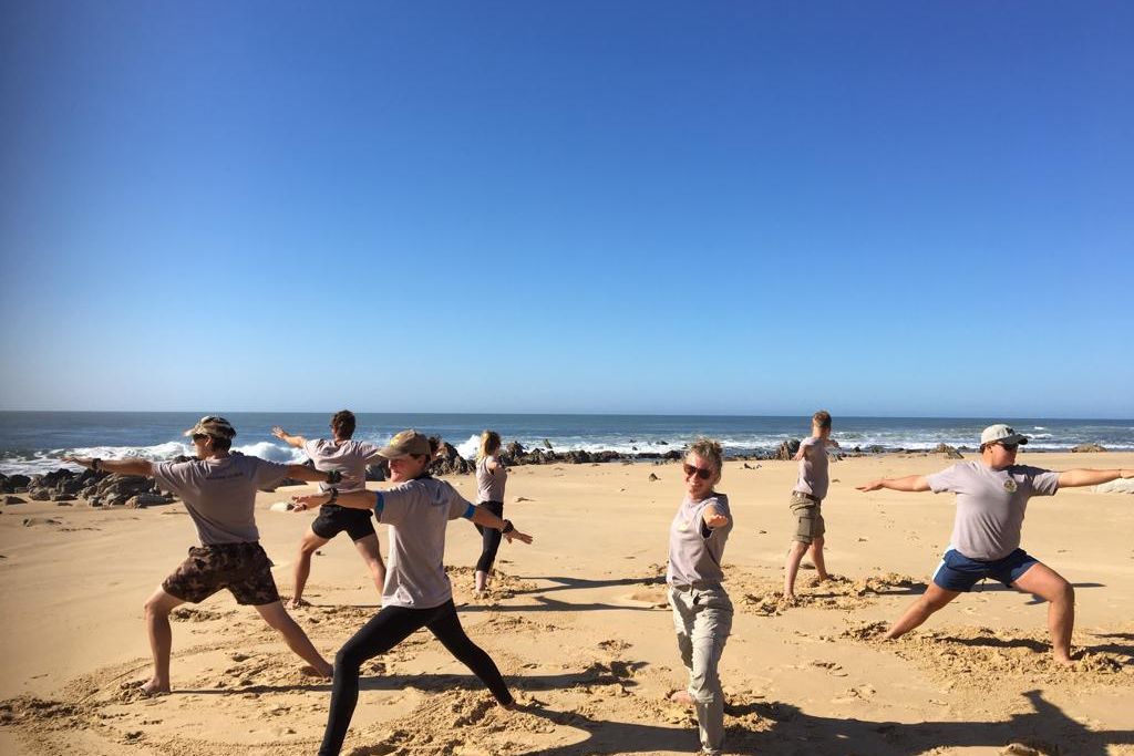 Marine Guide-Studenten stehen am Strand von Suedafrikas Sunshine Coast und machen gemeinsam Yoga