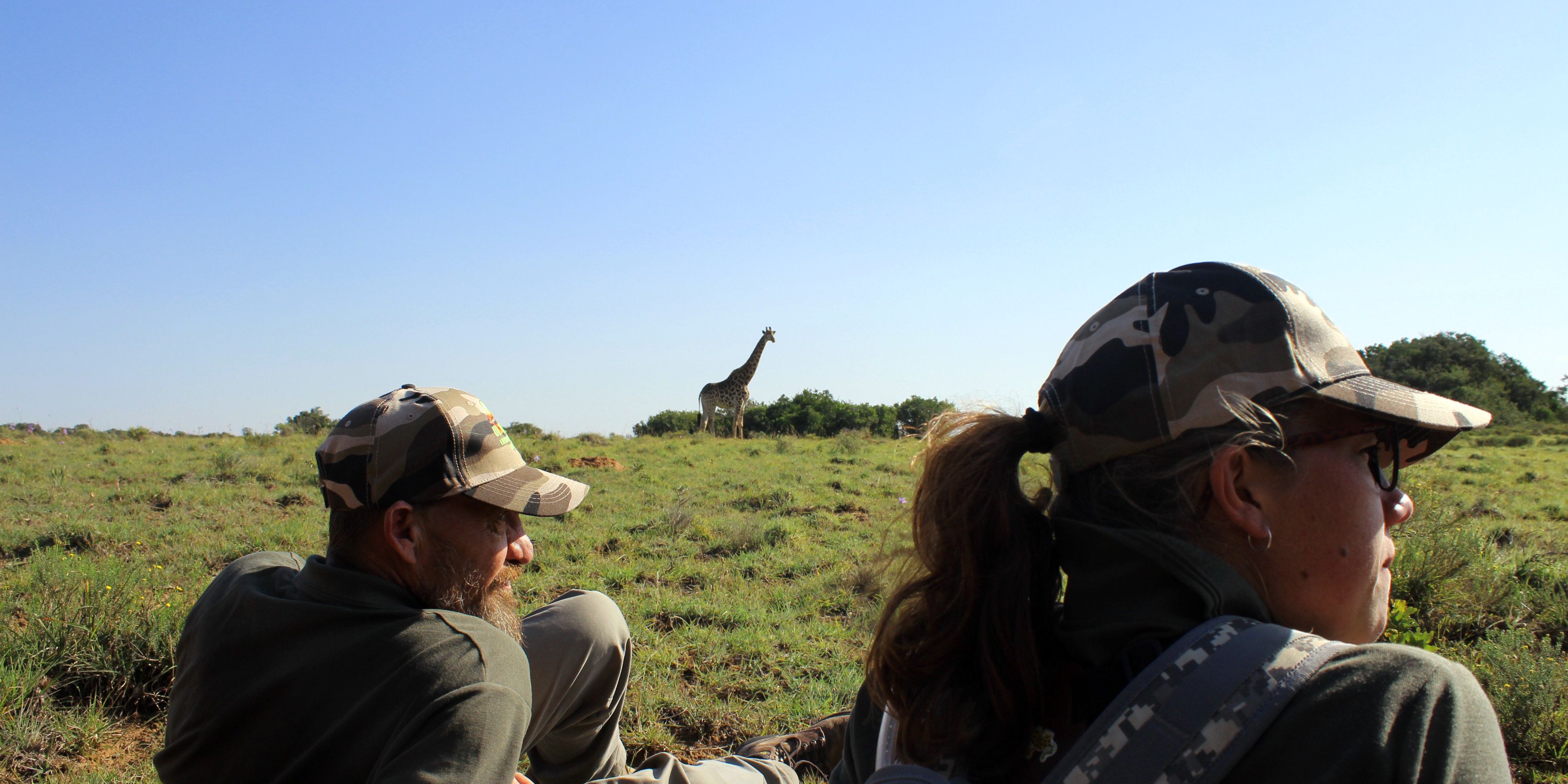Zwei Teilnehmer des Trails Guide-Kurses in Suedafrika liegen im Gras der Savanne; im Hintergrund ist eine Giraffe zu sehen