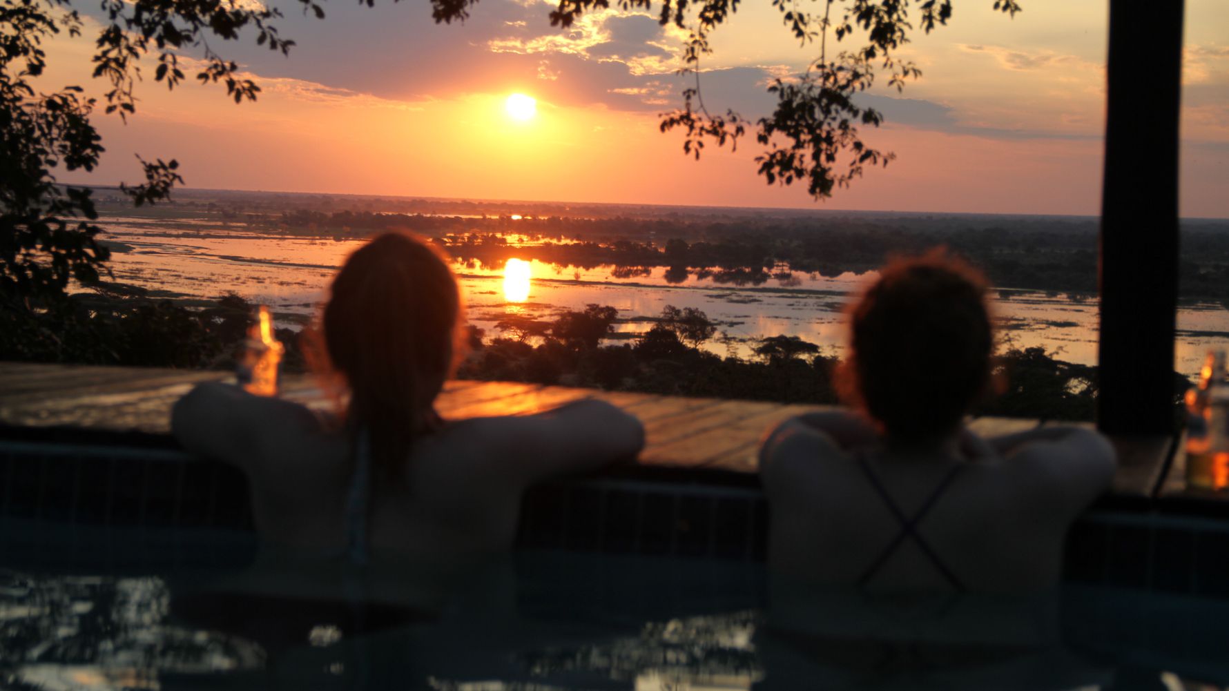 Luxus-Safari Afrika: Safari-Gaeste im Swimmingpool blicken bei Sonnenuntergang auf die Chobe Riverfront