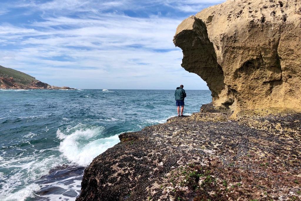 Aufnahme eines Marine Guide-Studenten, der an Kueste Suedafrikas auf einem Felsen steht und auf den Ozean blickt